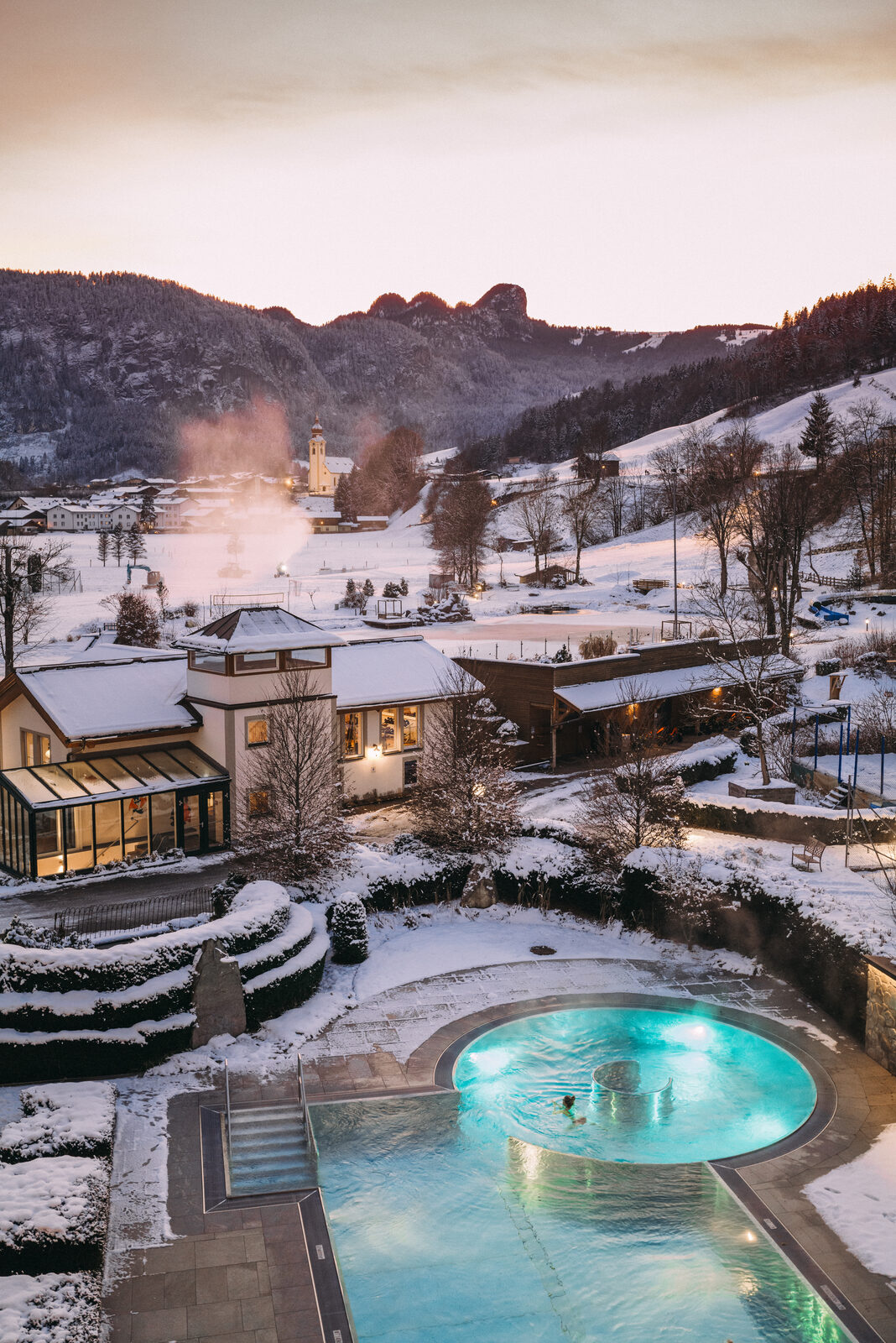 Aerial view of POST in winter — heated outdoor pool steaming, snow on the grounds, church and mountains behind.