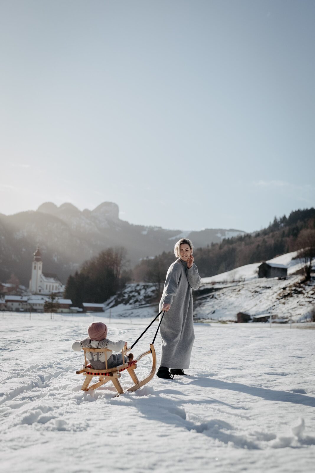 Mother pulls a child on a wooden sled across a snowfield in front of the village church.